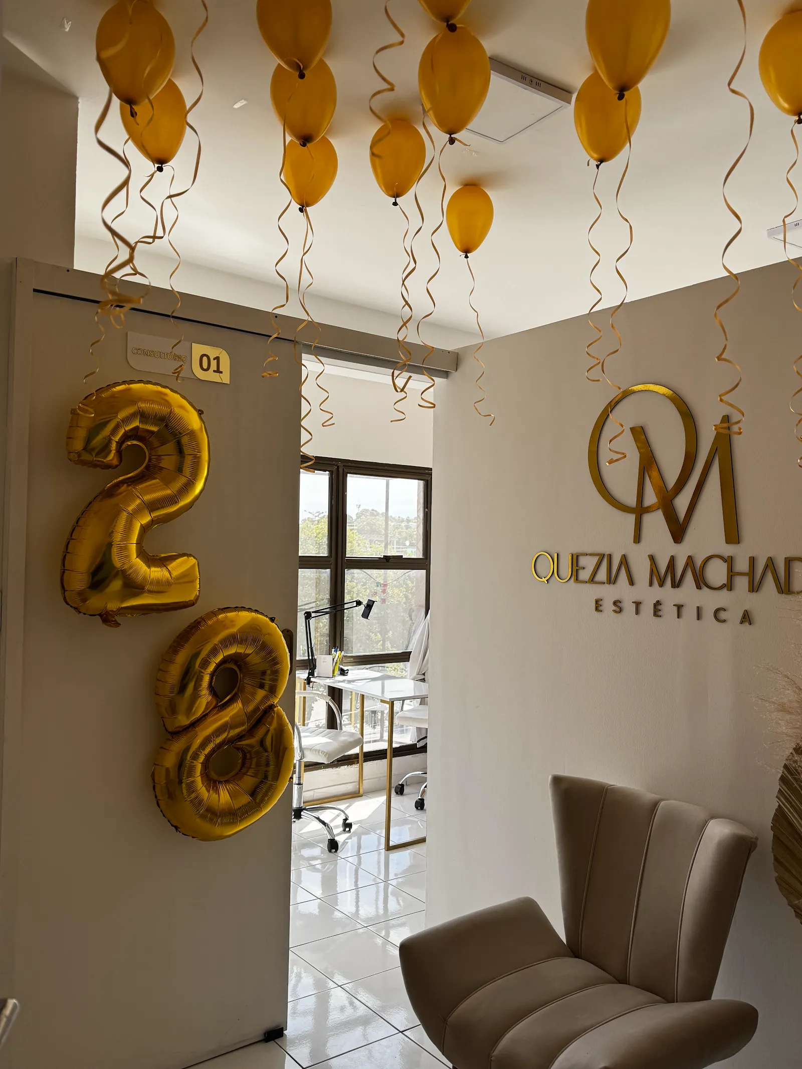 Recepção da clínica: parede clara com o logo dourado Quezia Machado Estética e uma poltrona de veludo bege.