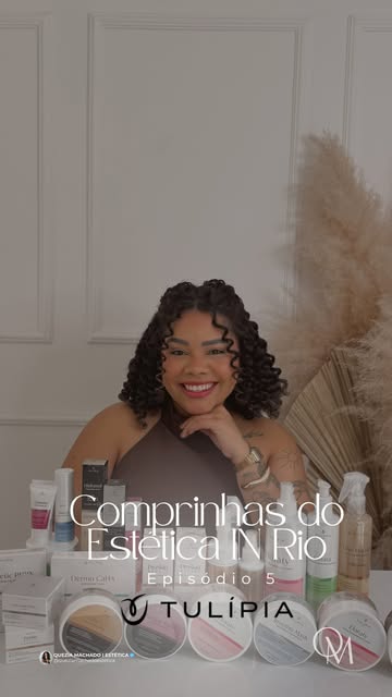 Quezia Machado sorrindo atrás de uma mesa com dermocosméticos, com capim-dos-pampas ao fundo.